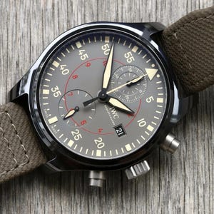 May include: A dark gray chronograph watch with a brown canvas strap. The dial features white Arabic numerals, luminous hands, a date window at the six o'clock position, and the text IWC SCHAFFHAUSEN. Two sub-dials are present on the face.
