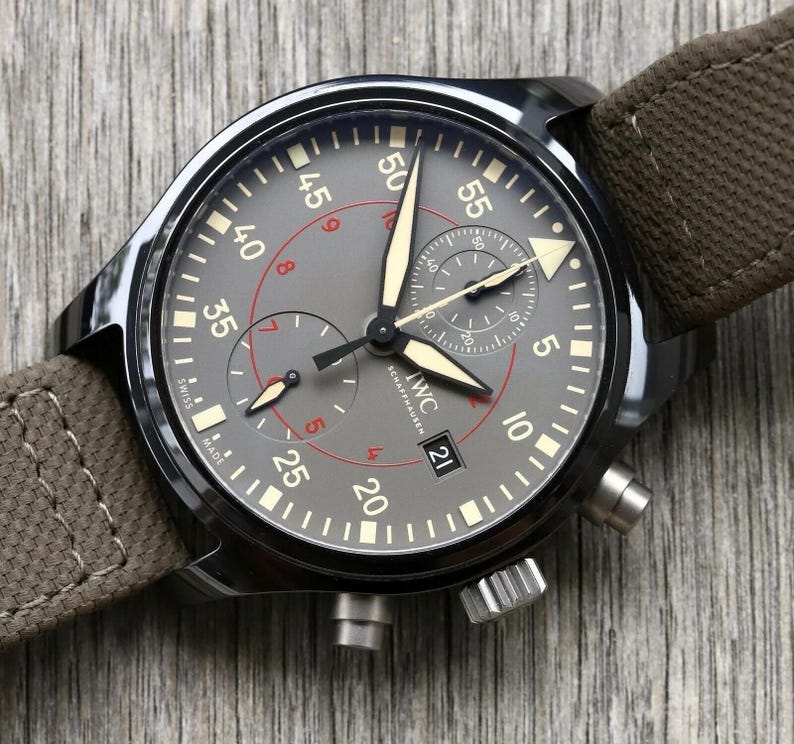 May include: A dark gray chronograph watch with a brown canvas strap. The dial features white Arabic numerals, luminous hands, a date window at the six o'clock position, and the text IWC SCHAFFHAUSEN. Two sub-dials are present on the face.