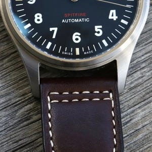 Puede incluir: Un reloj de pulsera con esfera negra que presenta n&uacute;meros ar&aacute;bigos blancos, el texto SPITFIRE AUTOMATIC en rojo y blanco, y SWISS MADE en la parte inferior. El reloj tiene una caja de metal plateado cepillado y una correa de cuero marr&oacute;n.