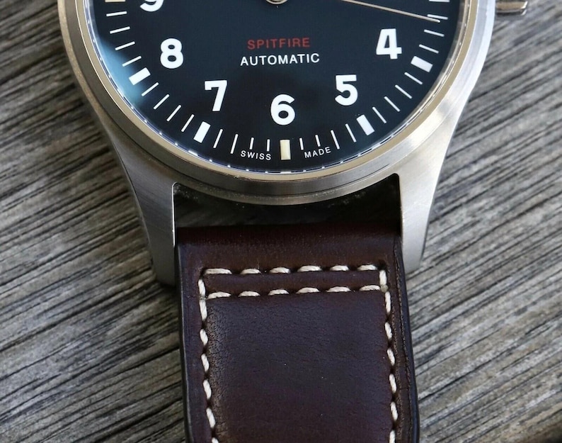 Puede incluir: Un reloj de pulsera con esfera negra que presenta n&uacute;meros ar&aacute;bigos blancos, el texto SPITFIRE AUTOMATIC en rojo y blanco, y SWISS MADE en la parte inferior. El reloj tiene una caja de metal plateado cepillado y una correa de cuero marr&oacute;n.
