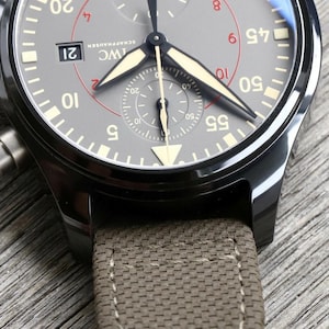 May include: An IWC Schaffhausen chronograph watch with a dark gray dial, luminous hands, and a date window at the three o'clock position. The watch features a black metal case and a textured olive green fabric strap with visible stitching.