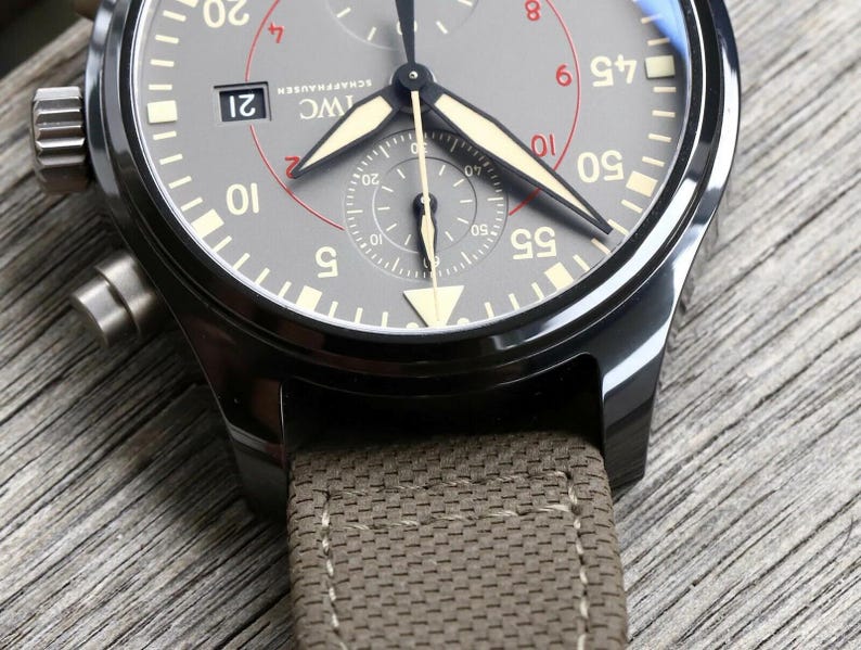 May include: An IWC Schaffhausen chronograph watch with a dark gray dial, luminous hands, and a date window at the three o'clock position. The watch features a black metal case and a textured olive green fabric strap with visible stitching.