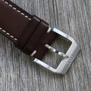 Puede incluir: Una correa de reloj de cuero marr&oacute;n oscuro con costuras blancas en contraste y una hebilla de espiga de metal plateado cepillado. La hebilla est&aacute; grabada con las letras IWC y la correa incluye dos trabillas de cuero para asegurar el extremo.