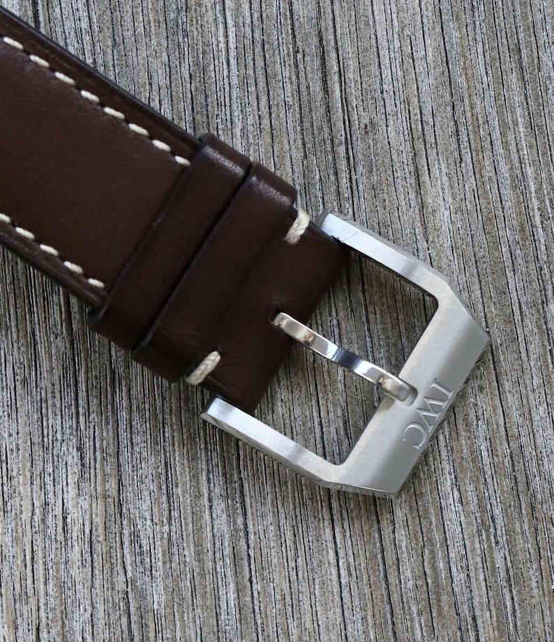 Puede incluir: Una correa de reloj de cuero marr&oacute;n oscuro con costuras blancas en contraste y una hebilla de espiga de metal plateado cepillado. La hebilla est&aacute; grabada con las letras IWC y la correa incluye dos trabillas de cuero para asegurar el extremo.