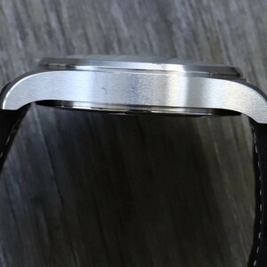 Puede incluir: Una vista de perfil de un reloj de pulsera con una caja de metal plateado cepillado y una correa de silicona negra con costuras blancas. La caja del reloj tiene un borde liso y redondeado y una parte superior plana, mostrando la uni&oacute;n entre la correa y las asas.