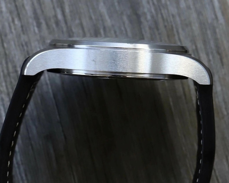 Puede incluir: Una vista de perfil de un reloj de pulsera con una caja de metal plateado cepillado y una correa de silicona negra con costuras blancas. La caja del reloj tiene un borde liso y redondeado y una parte superior plana, mostrando la uni&oacute;n entre la correa y las asas.