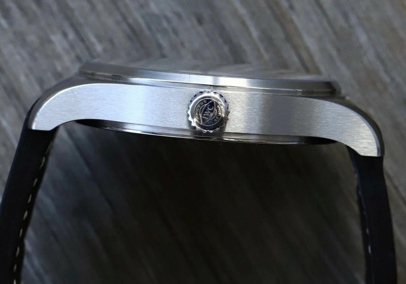 Puede incluir: Una vista lateral de una caja de reloj de acero inoxidable en tono plateado con acabado cepillado. El reloj presenta una corona circular grabada con el logotipo de IWC y est&aacute; sujeto a una correa de caucho negra con costuras blancas visibles.