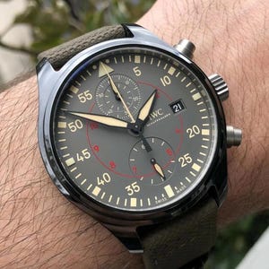 May include: A dark grey IWC Schaffhausen chronograph watch with a textured olive green strap. The dial features luminous hands, a date window at the three o'clock position, and a red inner circle with Arabic numerals. The watch has a metallic case.