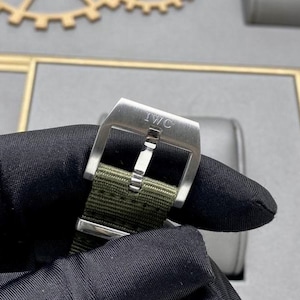 May include: A stainless steel watch buckle with the engraved text IWC, featuring a brushed metal finish and a central pin, attached to a textured olive green fabric watch strap with a matching metal keeper loop.