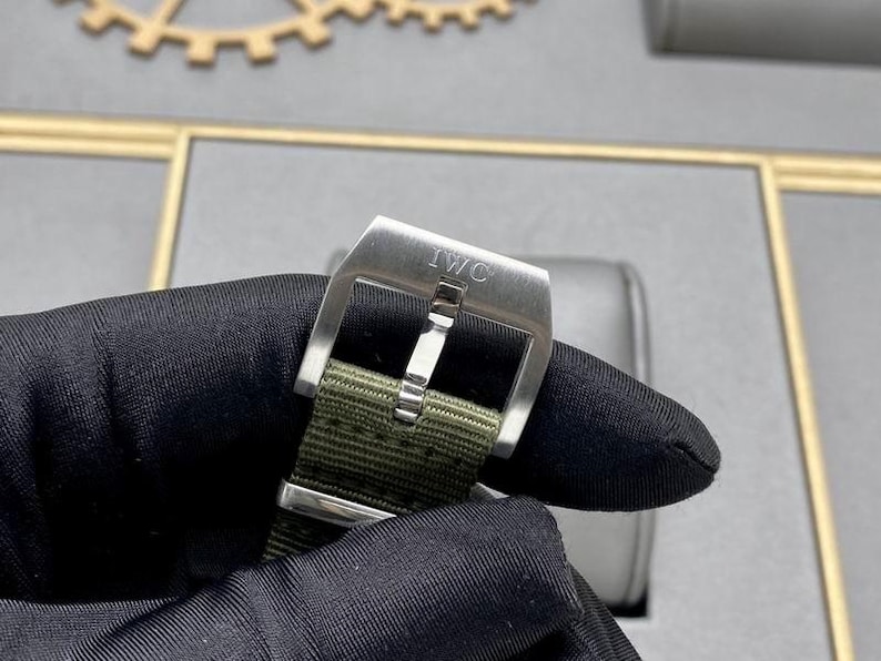 May include: A stainless steel watch buckle with the engraved text IWC, featuring a brushed metal finish and a central pin, attached to a textured olive green fabric watch strap with a matching metal keeper loop.