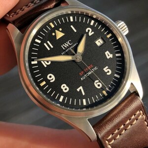 Puede incluir: Un reloj de pulsera autom&aacute;tico IWC Schaffhausen Spitfire con esfera negra, n&uacute;meros ar&aacute;bigos blancos, manecillas luminosas y una ventana de fecha en la posici&oacute;n de las tres. El reloj cuenta con una caja de acero inoxidable cepillado y una correa de cuero marr&oacute;n.