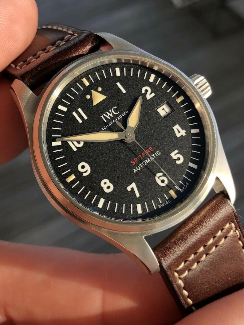 Puede incluir: Un reloj de pulsera autom&aacute;tico IWC Schaffhausen Spitfire con esfera negra, n&uacute;meros ar&aacute;bigos blancos, manecillas luminosas y una ventana de fecha en la posici&oacute;n de las tres. El reloj cuenta con una caja de acero inoxidable cepillado y una correa de cuero marr&oacute;n.