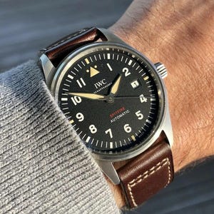 Puede incluir: Un reloj de pulsera IWC Schaffhausen Spitfire Automatic con caja redonda plateada, esfera negra con n&uacute;meros ar&aacute;bigos blancos, manecillas luminosas, ventana de fecha en la posici&oacute;n de las tres y correa de cuero marr&oacute;n con costuras blancas.