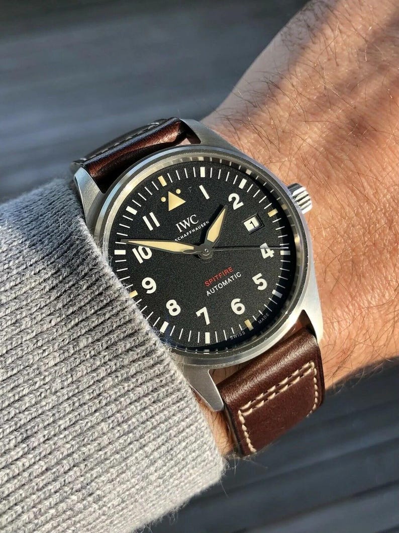 Puede incluir: Un reloj de pulsera IWC Schaffhausen Spitfire Automatic con caja redonda plateada, esfera negra con n&uacute;meros ar&aacute;bigos blancos, manecillas luminosas, ventana de fecha en la posici&oacute;n de las tres y correa de cuero marr&oacute;n con costuras blancas.