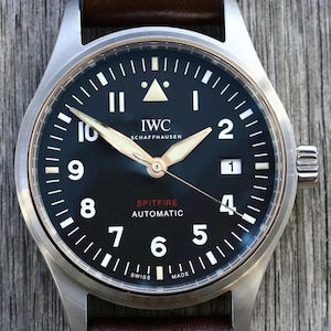 Puede incluir: Un reloj autom&aacute;tico IWC Schaffhausen Spitfire con esfera negra, n&uacute;meros ar&aacute;bigos blancos y ventana de fecha en la posici&oacute;n de las tres. El reloj cuenta con manecillas luminosas, un marcador triangular a las doce y una correa de cuero marr&oacute;n.