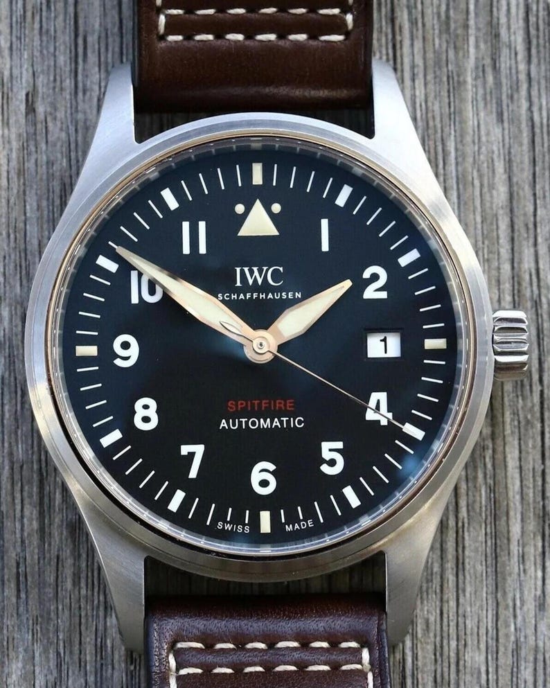 Puede incluir: Un reloj autom&aacute;tico IWC Schaffhausen Spitfire con esfera negra, n&uacute;meros ar&aacute;bigos blancos y ventana de fecha en la posici&oacute;n de las tres. El reloj cuenta con manecillas luminosas, un marcador triangular a las doce y una correa de cuero marr&oacute;n.