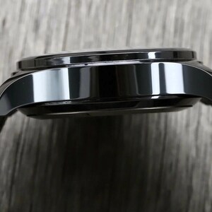 May include: A side profile view of a polished black watch case with a smooth, reflective metallic finish. The watch features curved lugs extending from the main body and is attached to a textured, brown fabric strap with a visible woven pattern.
