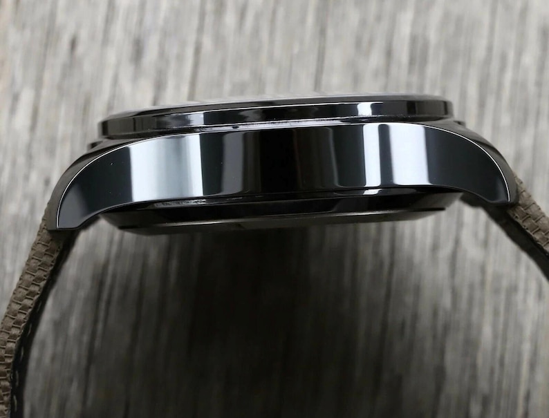 May include: A side profile view of a polished black watch case with a smooth, reflective metallic finish. The watch features curved lugs extending from the main body and is attached to a textured, brown fabric strap with a visible woven pattern.