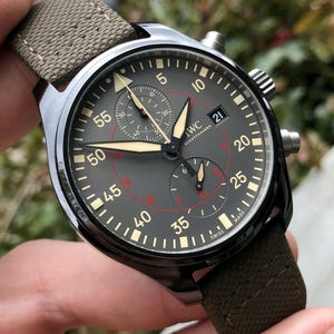 May include: An IWC Schaffhausen chronograph watch with a dark gray dial, luminous hands, and a textured olive green fabric strap. The dial features white Arabic numerals, a date window at the three o'clock position, and red inner markings.