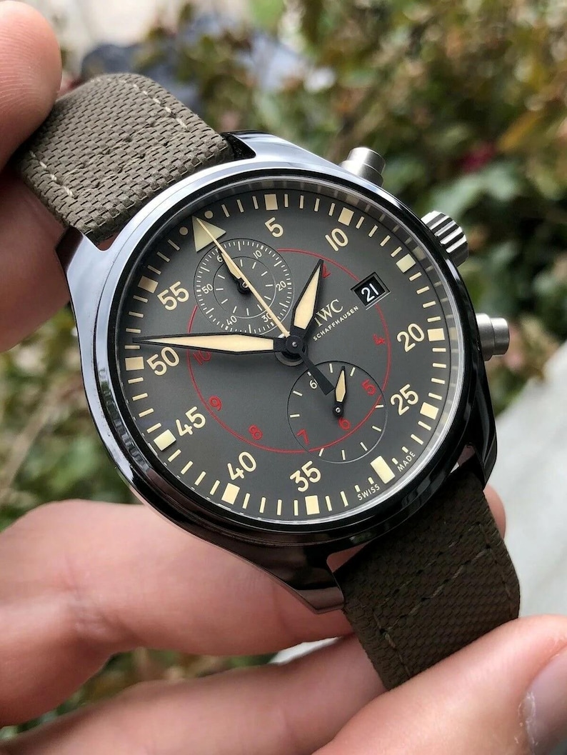 May include: An IWC Schaffhausen chronograph watch with a dark gray dial, luminous hands, and a textured olive green fabric strap. The dial features white Arabic numerals, a date window at the three o'clock position, and red inner markings.