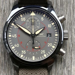 May include: A black ceramic chronograph watch with a grey dial featuring white and red numerals, luminous hands, and a date window at the three o'clock position. The watch face displays the IWC Schaffhausen logo and has a textured brown strap.