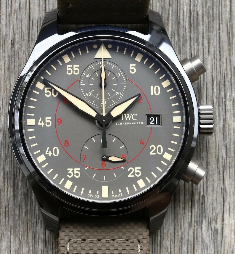 May include: A black ceramic chronograph watch with a grey dial featuring white and red numerals, luminous hands, and a date window at the three o'clock position. The watch face displays the IWC Schaffhausen logo and has a textured brown strap.