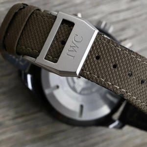 May include: A wristwatch featuring a textured olive green fabric strap with visible stitching and a brushed silver metal buckle engraved with the letters IWC. The watch case has a circular silver back with thin markers and a dark outer rim.