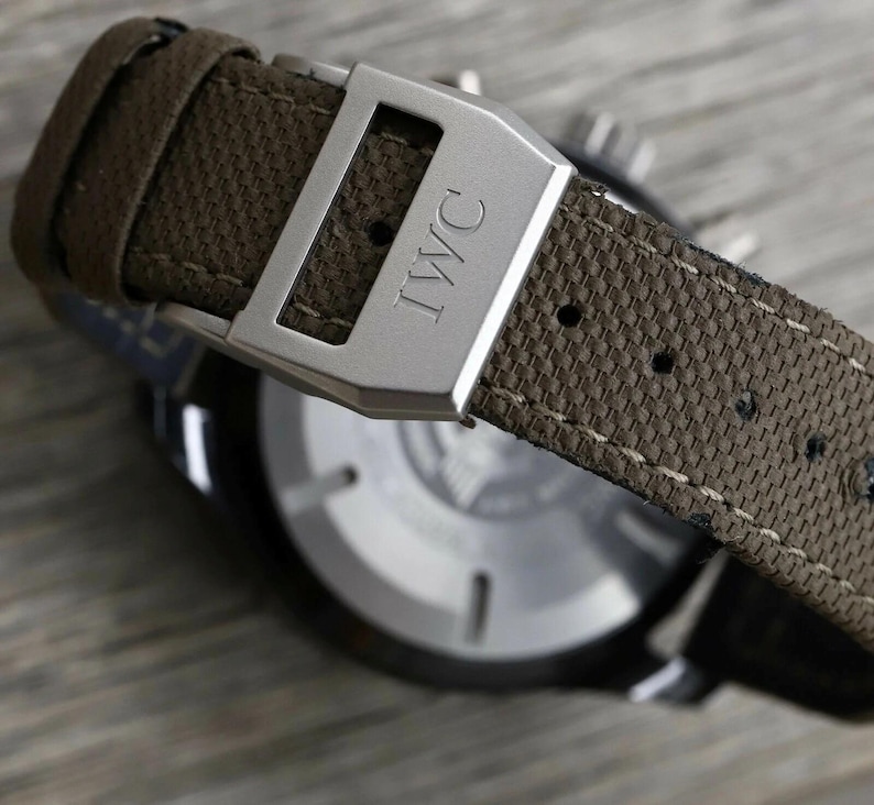 May include: A wristwatch featuring a textured olive green fabric strap with visible stitching and a brushed silver metal buckle engraved with the letters IWC. The watch case has a circular silver back with thin markers and a dark outer rim.