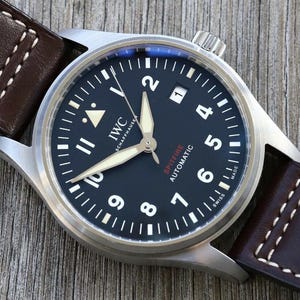 Puede incluir: Un reloj IWC Schaffhausen Spitfire Automatic con esfera negra, n&uacute;meros ar&aacute;bigos blancos y manecillas luminosas. El reloj cuenta con una ventana de fecha en la posici&oacute;n de las tres y una correa de cuero marr&oacute;n oscuro con costuras blancas.