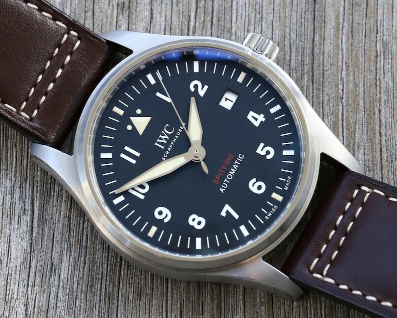 Puede incluir: Un reloj IWC Schaffhausen Spitfire Automatic con esfera negra, n&uacute;meros ar&aacute;bigos blancos y manecillas luminosas. El reloj cuenta con una ventana de fecha en la posici&oacute;n de las tres y una correa de cuero marr&oacute;n oscuro con costuras blancas.