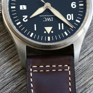 Puede incluir: Un reloj de pulsera de acero inoxidable con esfera negra mate, que presenta n&uacute;meros ar&aacute;bigos blancos en las posiciones de las dos, nueve y diez, una ventana de fecha, manecillas luminosas y una correa de cuero marr&oacute;n con costuras blancas.