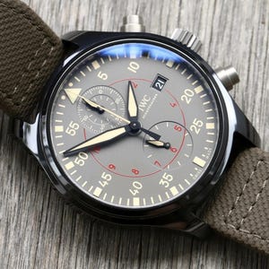 May include: A black chronograph wristwatch with a grey dial, featuring beige luminescent hands and markers, a date window at the three o'clock position, and a brown fabric strap. The dial displays the IWC Schaffhausen logo and Swiss Made text.