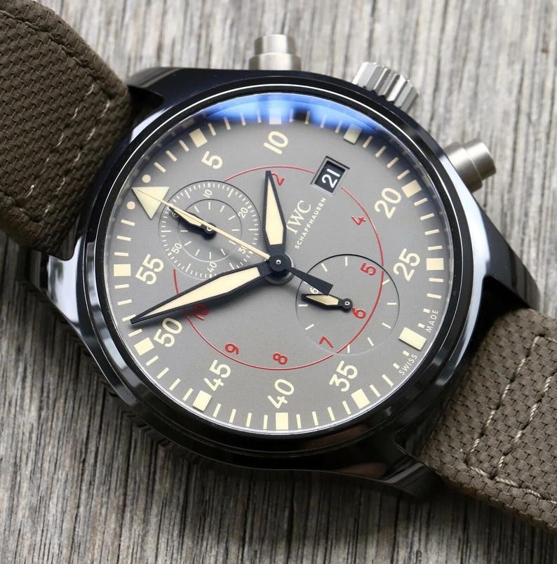 May include: A black chronograph wristwatch with a grey dial, featuring beige luminescent hands and markers, a date window at the three o'clock position, and a brown fabric strap. The dial displays the IWC Schaffhausen logo and Swiss Made text.