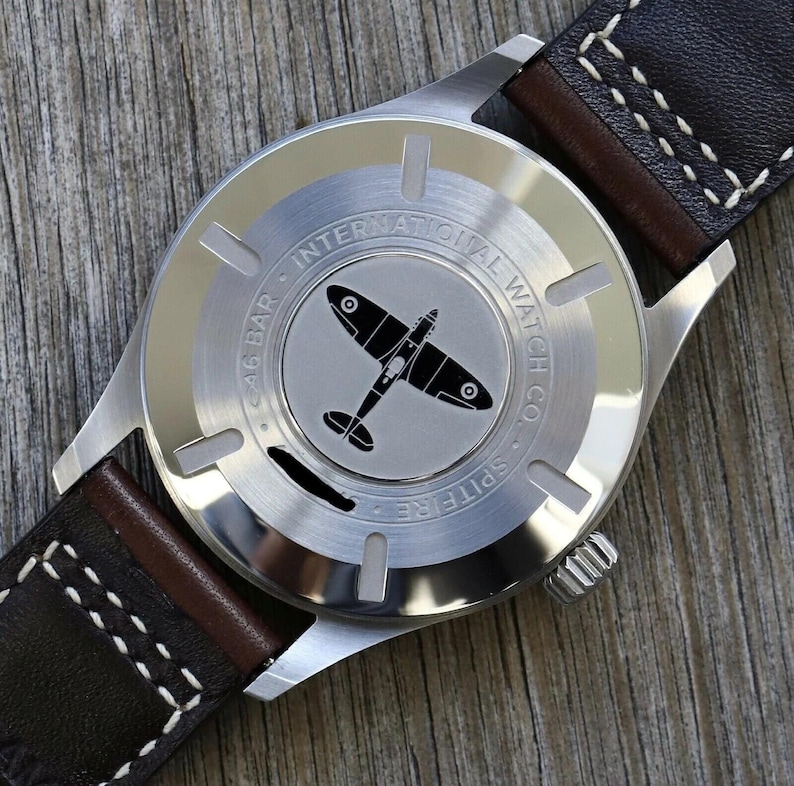 Puede incluir: Una caja de reloj de acero inoxidable con la silueta grabada de un avi&oacute;n Spitfire en el centro. El texto dice INTERNATIONAL WATCH CO. y 6 BAR. El reloj est&aacute; sujeto a una correa de cuero marr&oacute;n oscuro con costuras blancas en contraste.