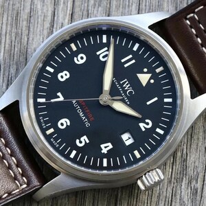 Puede incluir: Un reloj de pulsera autom&aacute;tico IWC Schaffhausen Spitfire con esfera negra, n&uacute;meros ar&aacute;bigos blancos, manecillas luminosas y una ventana de fecha en la posici&oacute;n de las tres. El reloj cuenta con una caja de acero inoxidable cepillado y una correa de cuero marr&oacute;n.