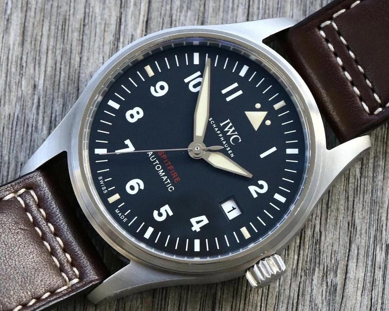 Puede incluir: Un reloj de pulsera autom&aacute;tico IWC Schaffhausen Spitfire con esfera negra, n&uacute;meros ar&aacute;bigos blancos, manecillas luminosas y una ventana de fecha en la posici&oacute;n de las tres. El reloj cuenta con una caja de acero inoxidable cepillado y una correa de cuero marr&oacute;n.