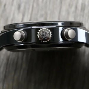 May include: A black chronograph watch case with a side profile view. The case features a central crown embossed with the IWC logo and two silver-toned pushers on either side. A textured brown strap is attached to the lugs of the watch.