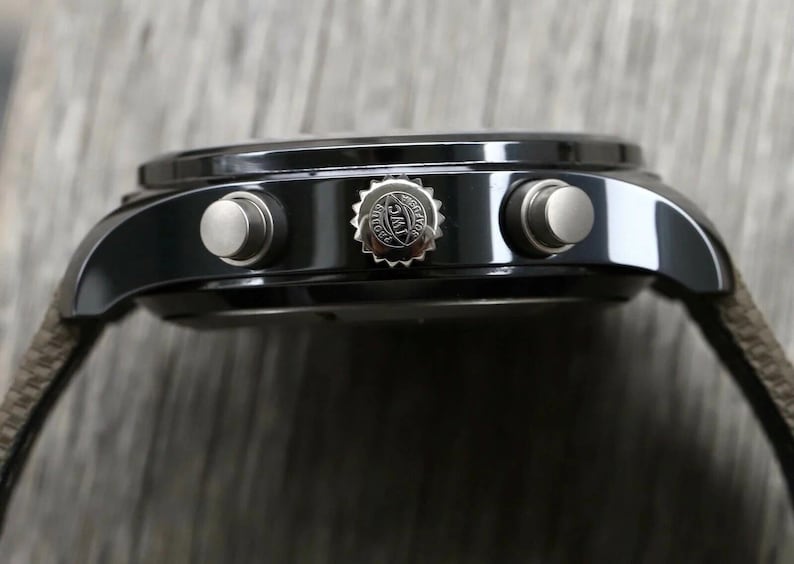 May include: A black chronograph watch case with a side profile view. The case features a central crown embossed with the IWC logo and two silver-toned pushers on either side. A textured brown strap is attached to the lugs of the watch.