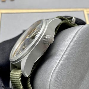 May include: A silver-toned wristwatch with a dark dial and a textured olive green fabric strap. The watch features a round brushed metal case, a side-mounted winding crown with an embossed logo, and a clear crystal face showing hour markers.
