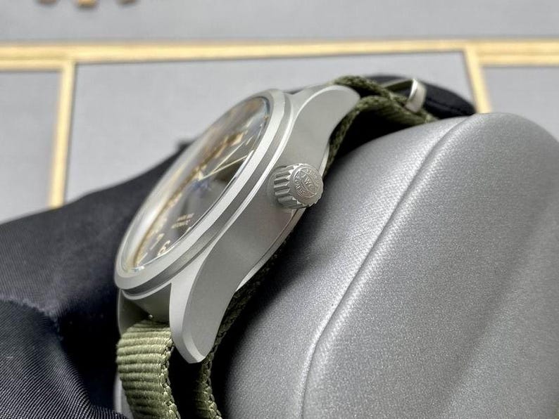 May include: A silver-toned wristwatch with a dark dial and a textured olive green fabric strap. The watch features a round brushed metal case, a side-mounted winding crown with an embossed logo, and a clear crystal face showing hour markers.