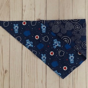 Może przedstawiać: Dwustronna chusta dla psa z granatowym wzorem Care Bears z chmurami i sercami po jednej stronie oraz granatowym okrągłym wzorem po drugiej stronie. Chusta ma kształt trójkąta i jest wykonana z tkaniny.