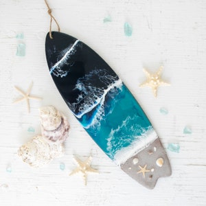 May include: A surfboard-shaped resin art piece with a blue and black ocean wave design. The piece has a white sandy beach at the bottom with seashells and a starfish.