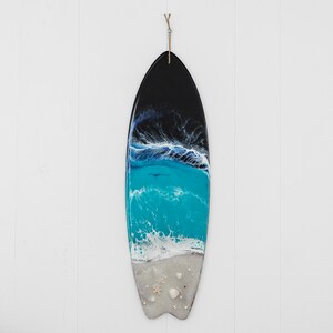 May include: A black and blue surfboard with a white foamy wave and seashells. The surfboard is hanging on a white wall.