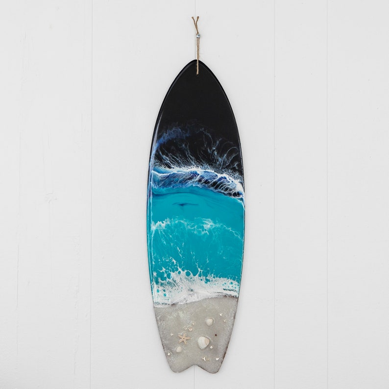 May include: A black and blue surfboard with a white foamy wave and seashells. The surfboard is hanging on a white wall.