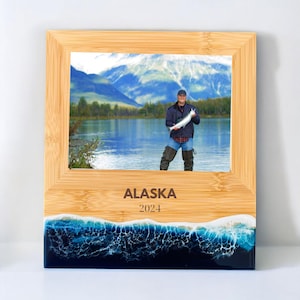 May include: A wooden picture frame with a blue and white resin base. The frame has a photo of a person holding a fish in front of a lake and mountains. The frame is engraved with the text "ALASKA 2024".