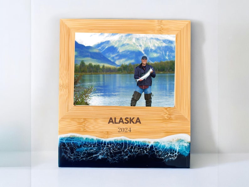 May include: A wooden picture frame with a blue and white resin base. The frame has a photo of a person holding a fish in front of a lake and mountains. The frame is engraved with the text "ALASKA 2024".