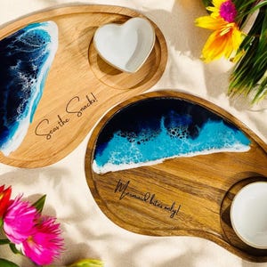 May include: Two wooden serving trays with blue and white resin ocean wave designs. The smaller tray has the text "Mermaid bites only!" and the larger tray has the text "Sea the Snacks!"