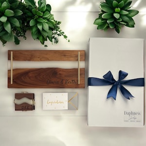 May include: A gift box set with a personalized wooden serving tray, a small wooden box with the initials "OK", a card that says "Congratulations", and a white gift box with a blue ribbon. The serving tray is engraved with the names "Olivia & Keith".