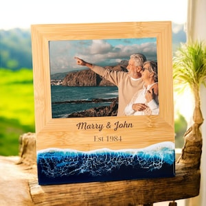May include: A wooden photo frame with a blue and white resin wave base. The frame has a photo of an older couple standing on a beach. The frame is engraved with the text "Marry & John Est 1984".