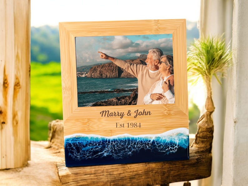 May include: A wooden photo frame with a blue and white resin wave base. The frame has a photo of an older couple standing on a beach. The frame is engraved with the text "Marry & John Est 1984".